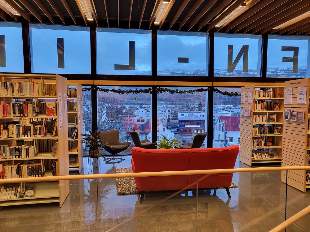 Mynd af bókahillum, sófa og glugga með útsýni yfir hluta af Akureyri. Jólaljós hanga yfir gluggunum