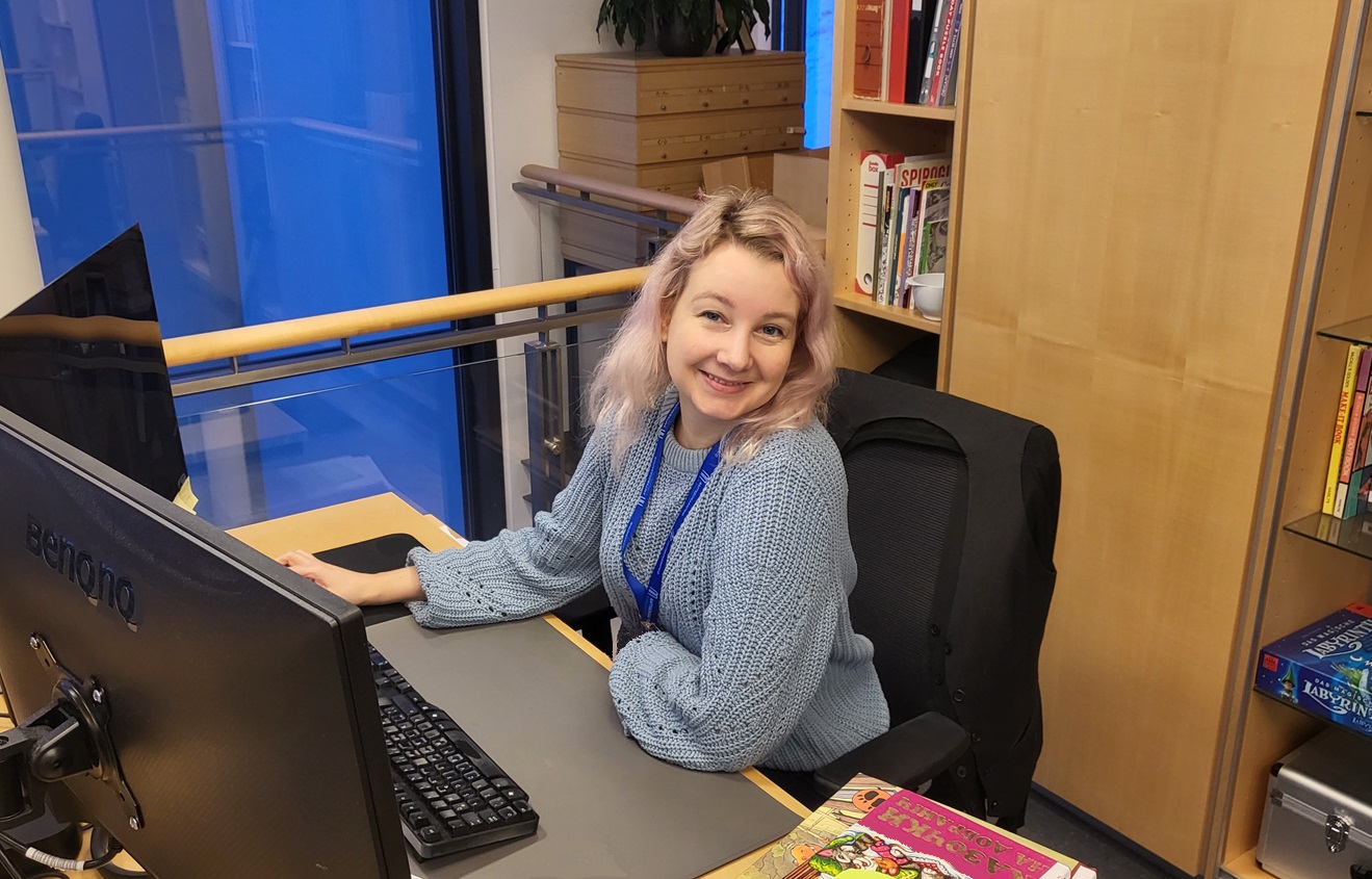 Woman sitting by a workdesk with computer and screens in front of her and shelves in the background