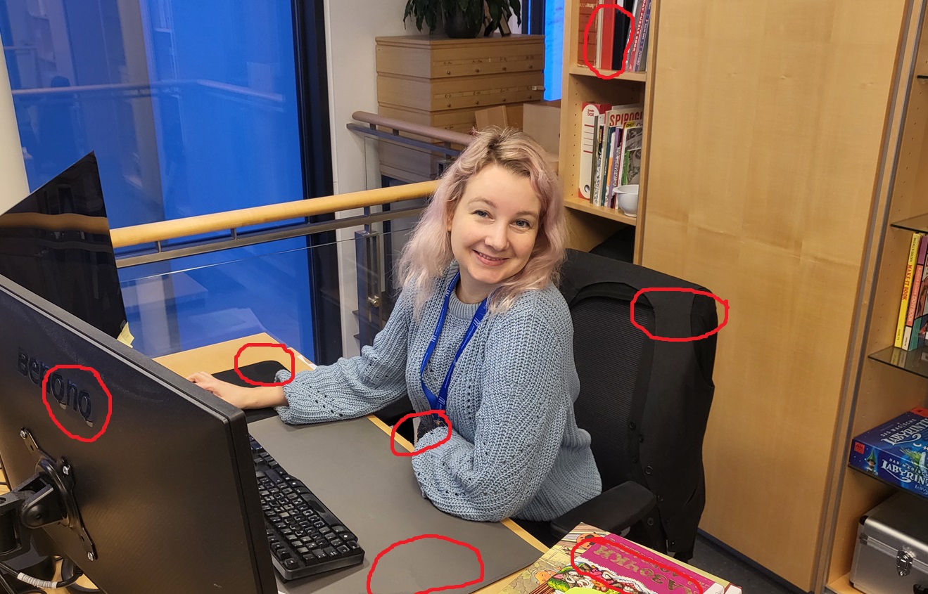 Kona við borð, tölva fyrir framan og skjáir, hillur fyrir aftan