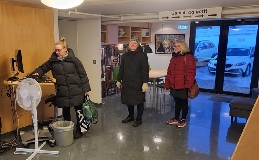 Three women logging in to work at the Municipal Library, computer on a desk and shelves with books in the background