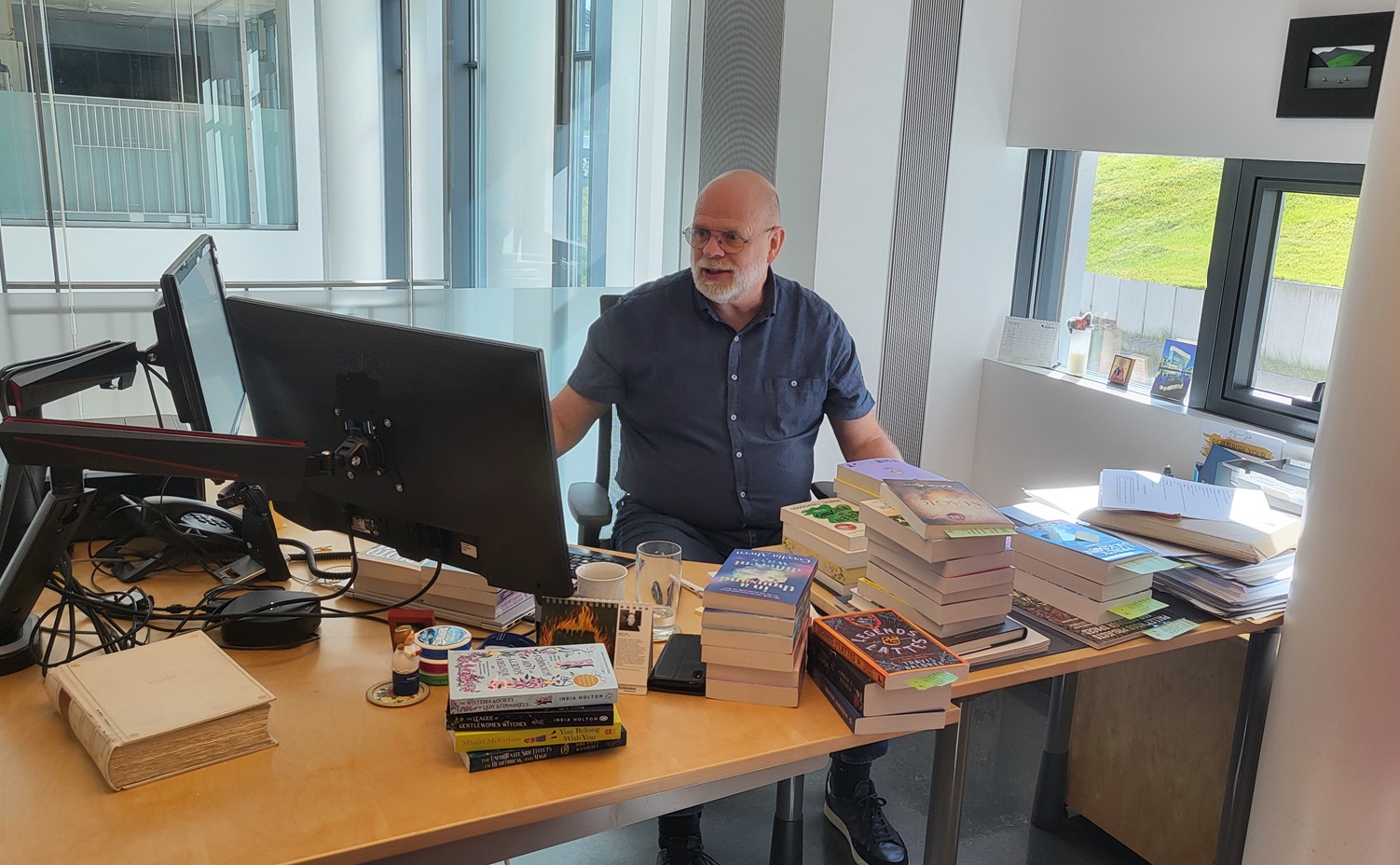 Man sitting behind a desk in front of a computer and lot of books lying on the desk